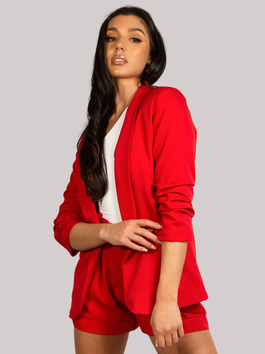 Female model stands in front of a plain background. The model wears a red open blazer with ruched 3/4 sleeves and a matching red shorts. The  model has tanned skin and long black hair, she faces the camera, placing one leg to the side, her arm across her stomach and one hand by her sode. The ruched sleeves can be seen. 