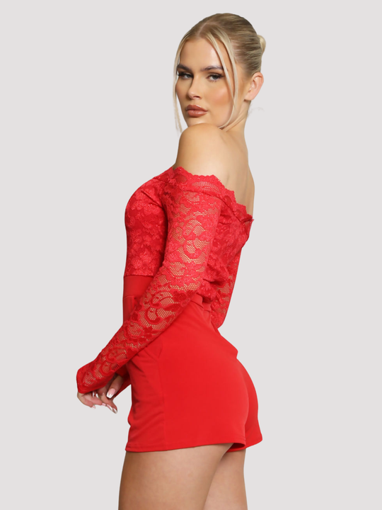 Female model stands in front of a plain background. The model wears a red lace playsuit, that has an off the shoulder bardot design and laced long sleeves and side pockets The model has blonde hair and tanned skin, she stands to the side and turns her head to look into the camera. The side of the red playsuit can be seen, including the off the shoulder lace design and laced sleeves.