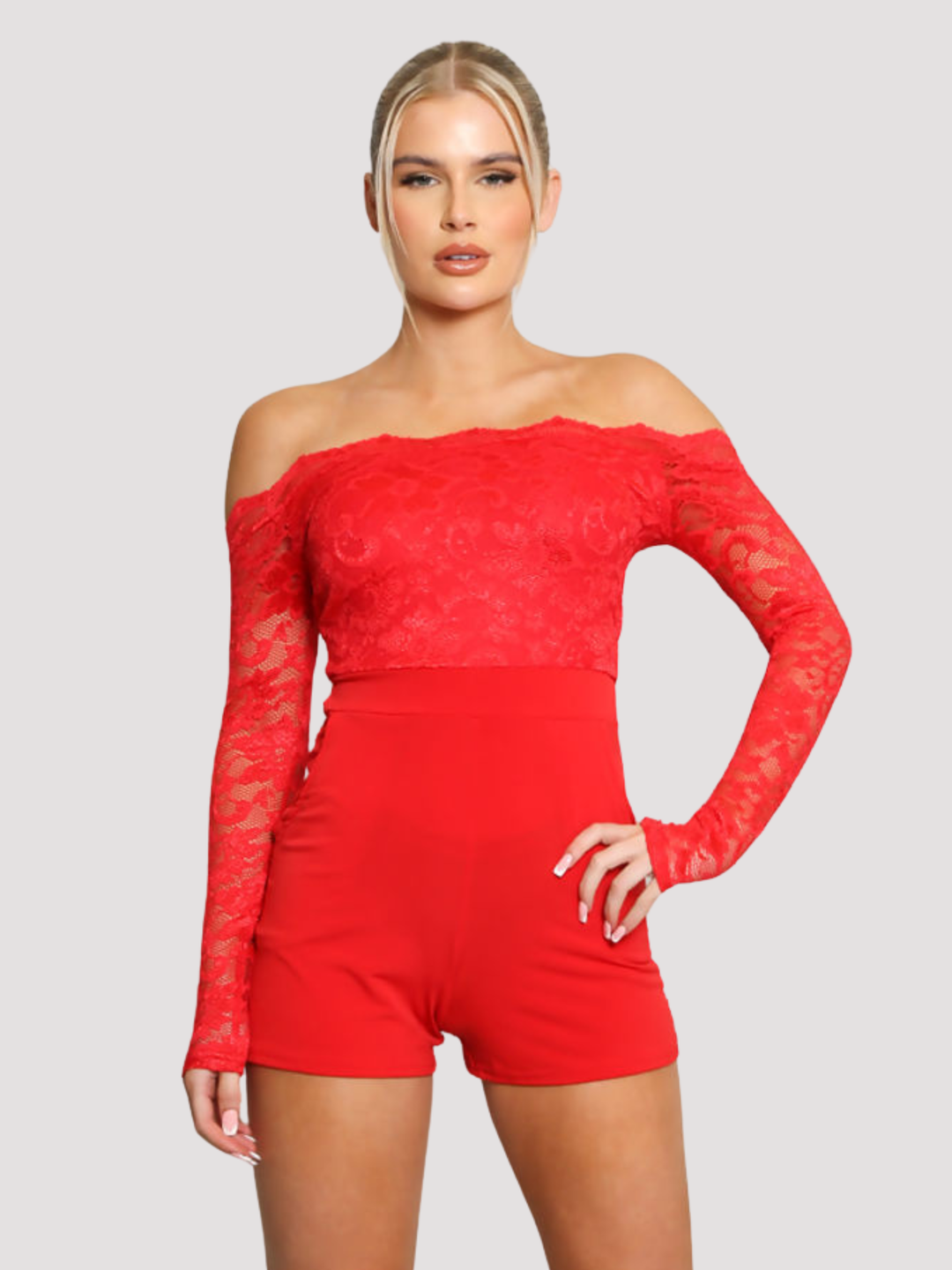 Female model stands in front of a plain background. The model wears a red lace playsuit, that has an off the shoulder bardot design and laced long sleeves and side pockets The model has blonde hair and tanned skin, she faces the camera and places one hand on her hip and the other by her side, the front of the lace playsuit can be seen. 