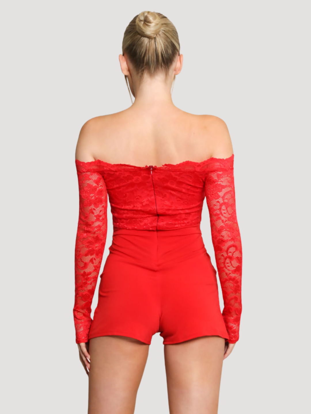 Female model stands in front of a plain background. The model wears a red lace playsuit, that has an off the shoulder bardot design and laced long sleeves and side pockets The model has blonde hair and tanned skin, she stands with her back to the camera, placing both hands by her sides. The back of the playsuit can be seen, including lace long sleeved, red laced playsuit and invisible zip back fastening. 