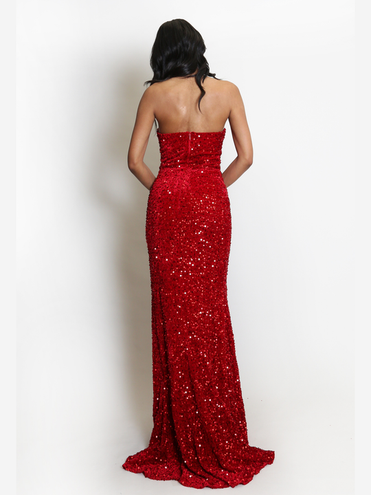 Full length shot. Model stands before a plain background. The model wears a red sequin maxi dress, with a fish tail hem and silver sequin sweetheart neckline. The model has light brown skin and long black hair. The model stands with her back to the camera, the long fish tail trail can be seen. 