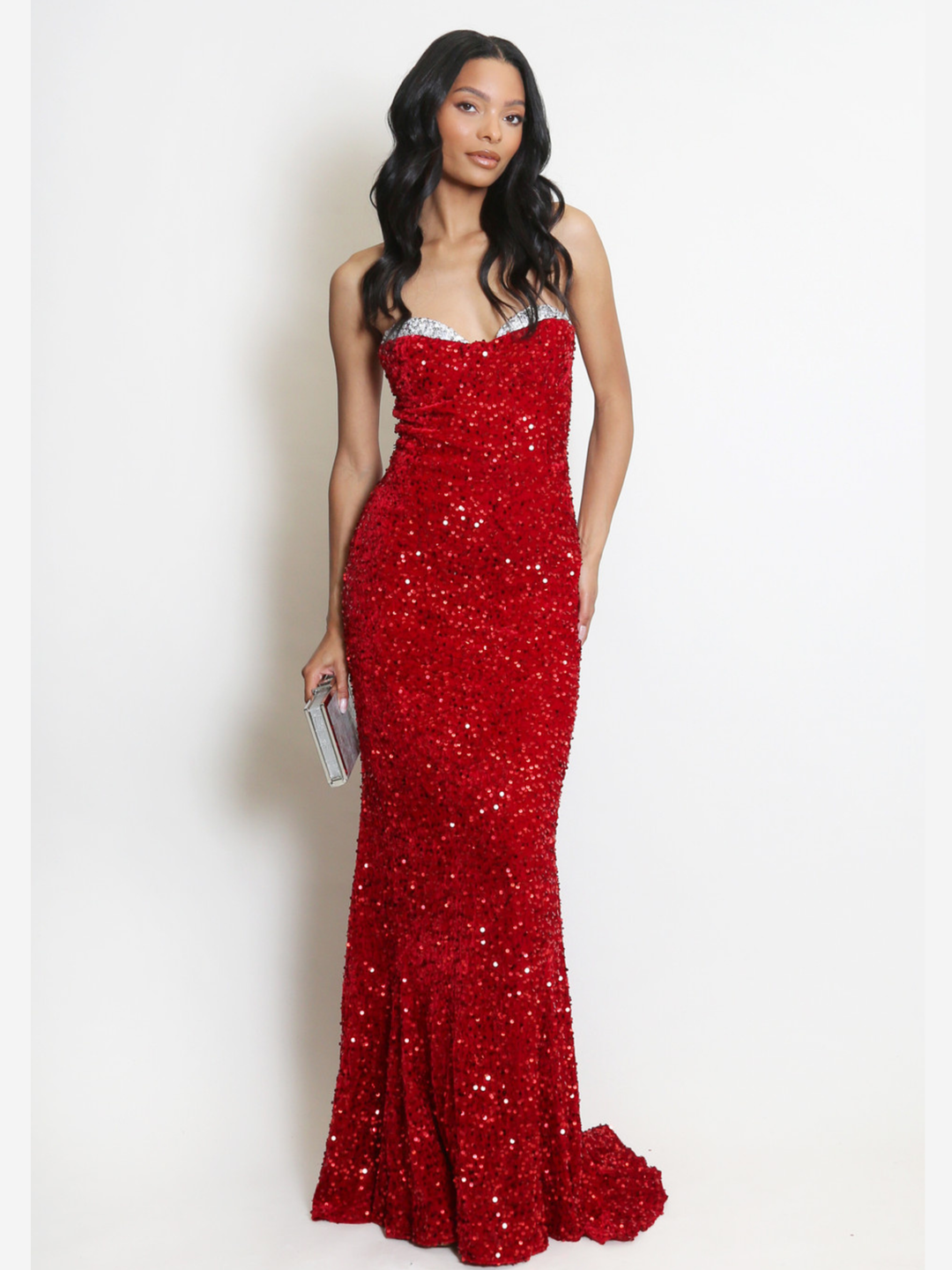 Full length shot. Model stands before a plain background. The model wears a red sequin maxi dress, with a fish tail hem and silver sequin sweetheart neckline. The model faces the camera, she has light brown skin and long black hair. The model  holds a Diamante clutch hand bag, the fish tail can be seen. 