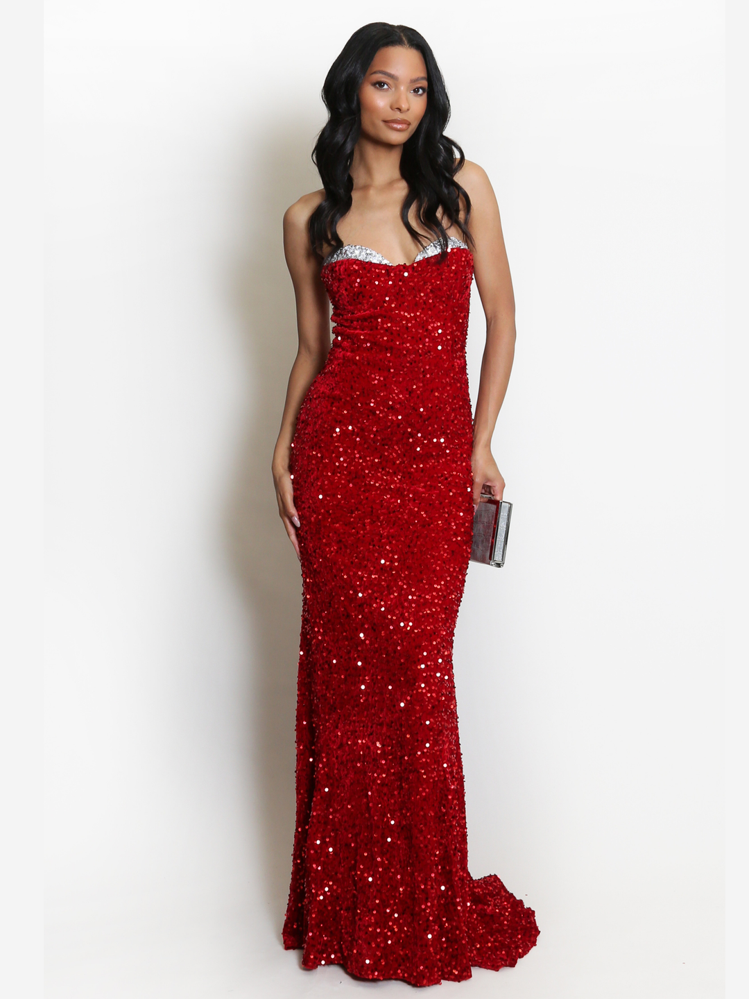 Full length shot. Model stands before a plain background. The model wears a red sequin maxi dress, with a fish tail hem and silver sequin sweetheart neckline. The model faces the camera, she has light brown skin and long black hair. The model faces the camera, she holds a Diamante clutch hand bag, the long fish tail can be seen.