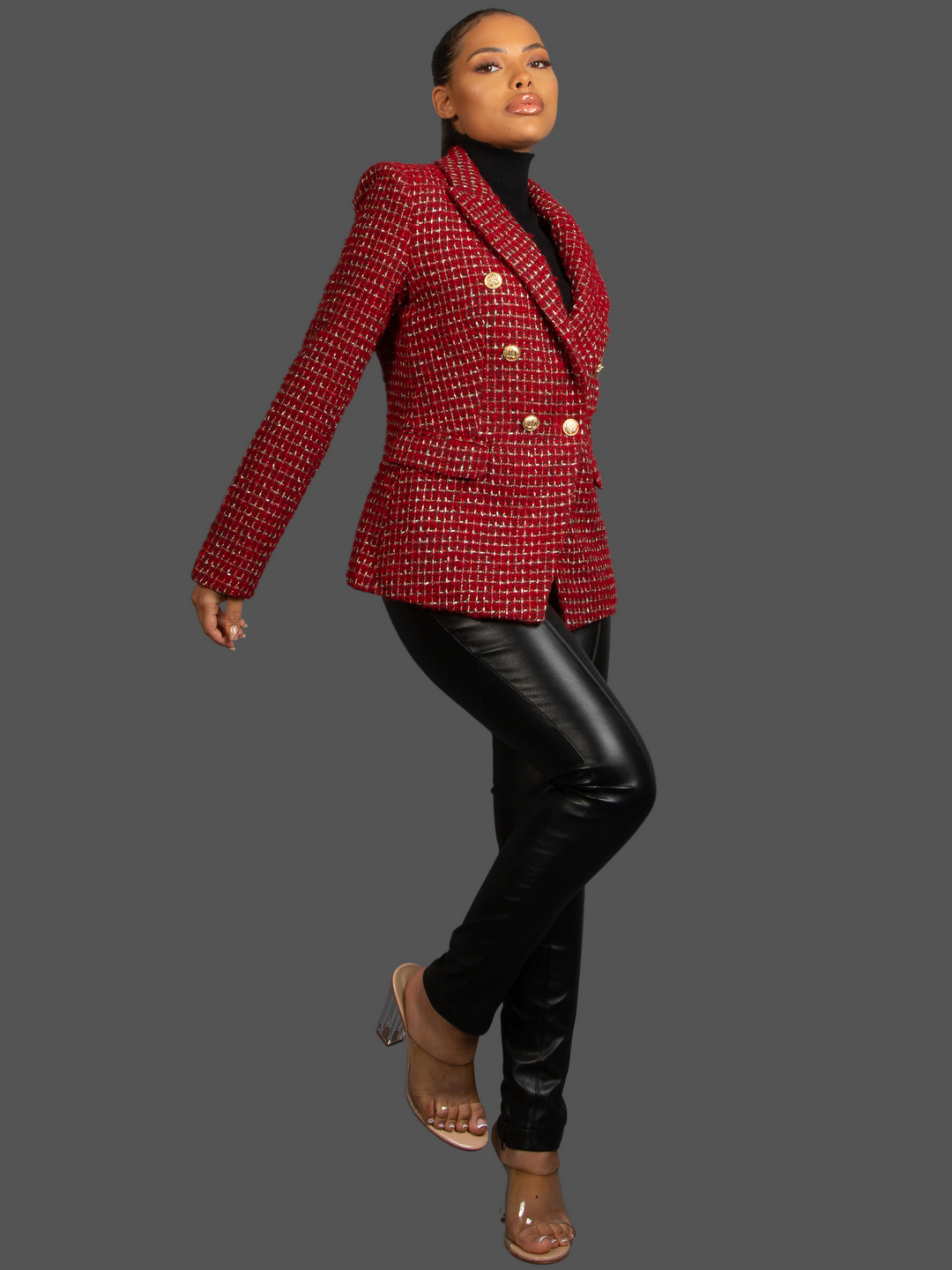 Full length shot. Model wears Red Wine Double Breasted Tweed Blazer, With Metallic Yarn Shimmer and Military Gold Buttons. The model as light brown skin and her hair pulled back into a ponytail. Model wears a faux leather trousers and eels, she bends one knee and stretches her arm out, the front and side of the blazer can be seen. 