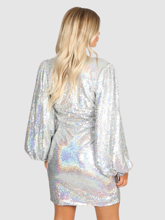 Model stands in front of a plain grey background. The model wears a silver sequin mini dress with a v cut  wrap front and puffy sleeves. The model  has tanned skin and blonde hair, the model stands with her back to the camera, the puffy sleeves can be seen.