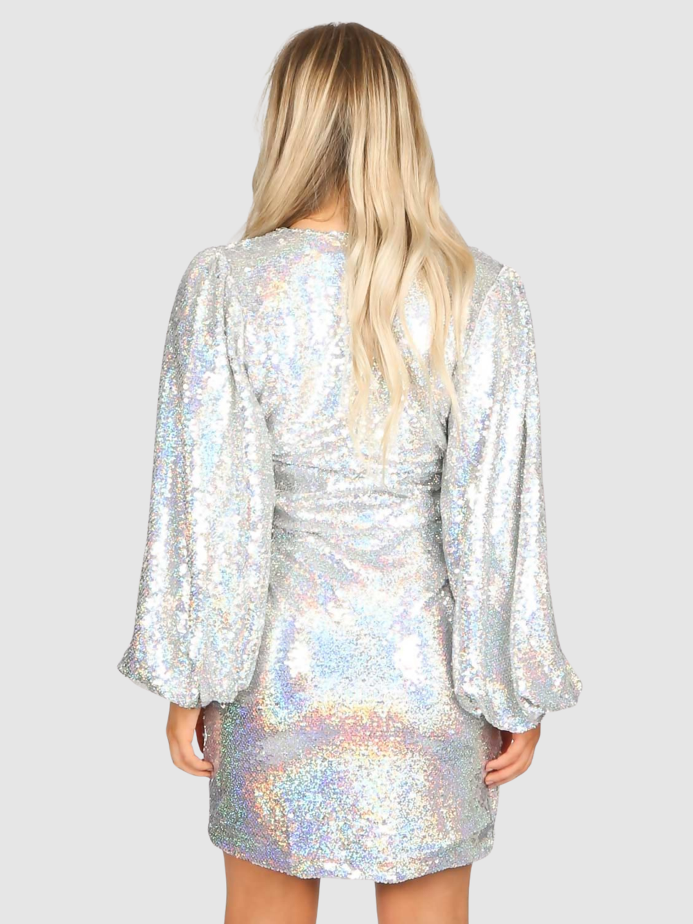 Model stands in front of a plain grey background. The model wears a silver sequin mini dress with a v cut  wrap front and puffy sleeves. The model  has tanned skin and blonde hair, the model stands with her back to the camera, the bck of her mini dress can be seen.
