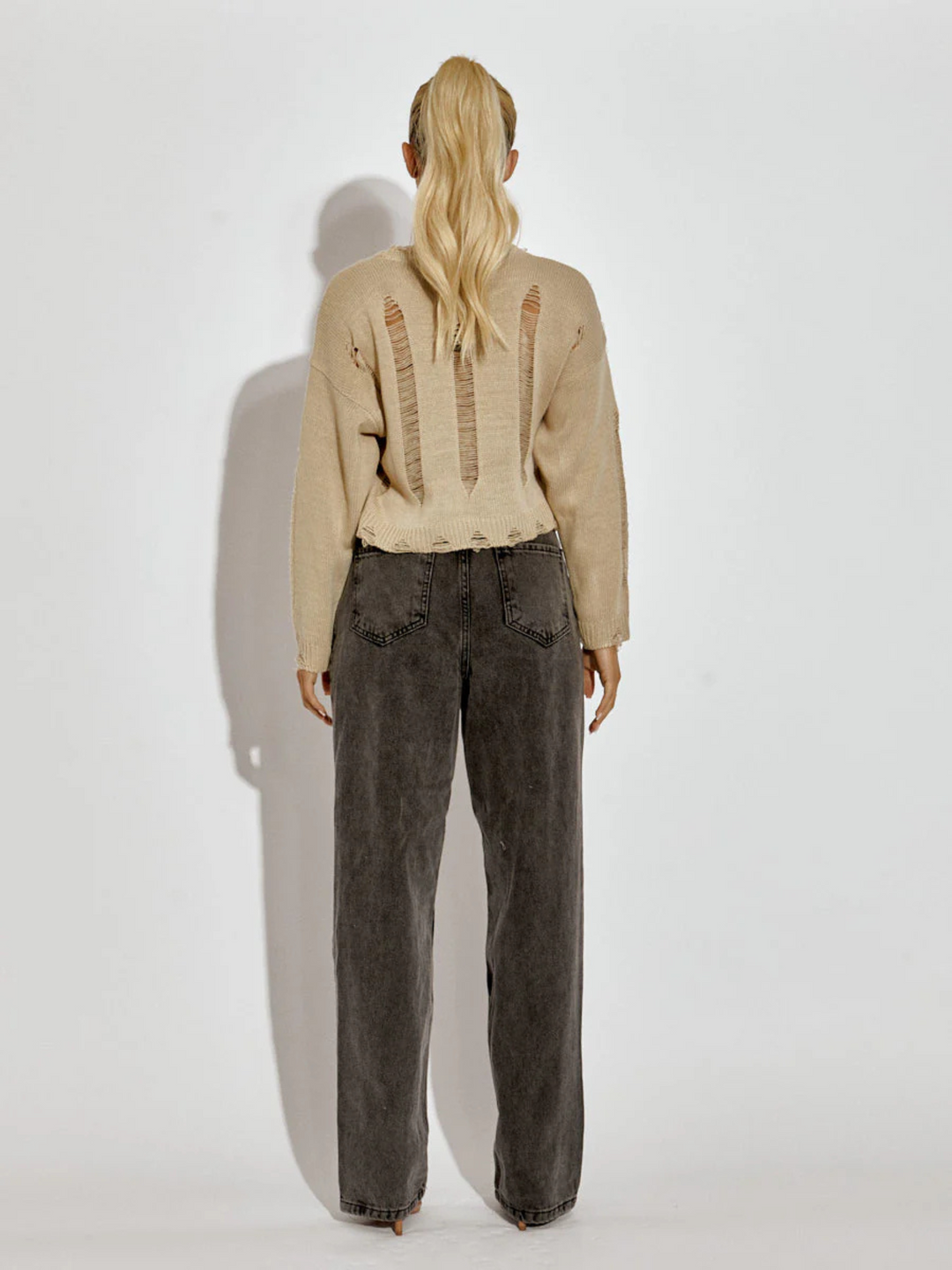 Full length shot. A female model wearing a stone long sleeve distressed style knitted cropped jumper in stone color paired with black jeans. The stone distressed knitted jumper has distressed panelling  and distressed hems. The model has tanned skin and long blonde hair that is in a ponytail. The model stands with her back to the camers, the back of the distressed knitted jumper can be seen.