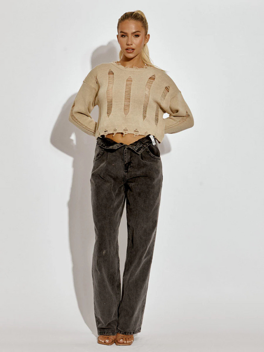 Full length shot. A female model wearing a stone long sleeve distressed style knitted cropped jumper in stone color paired with black jeans. The stone distressed knitted jumper has distressed panelling  and distressed hems. The model has tanned skin and long blonde hair that is in a ponytail. The model plaes her arms behind her back and stares directly into the camera. 