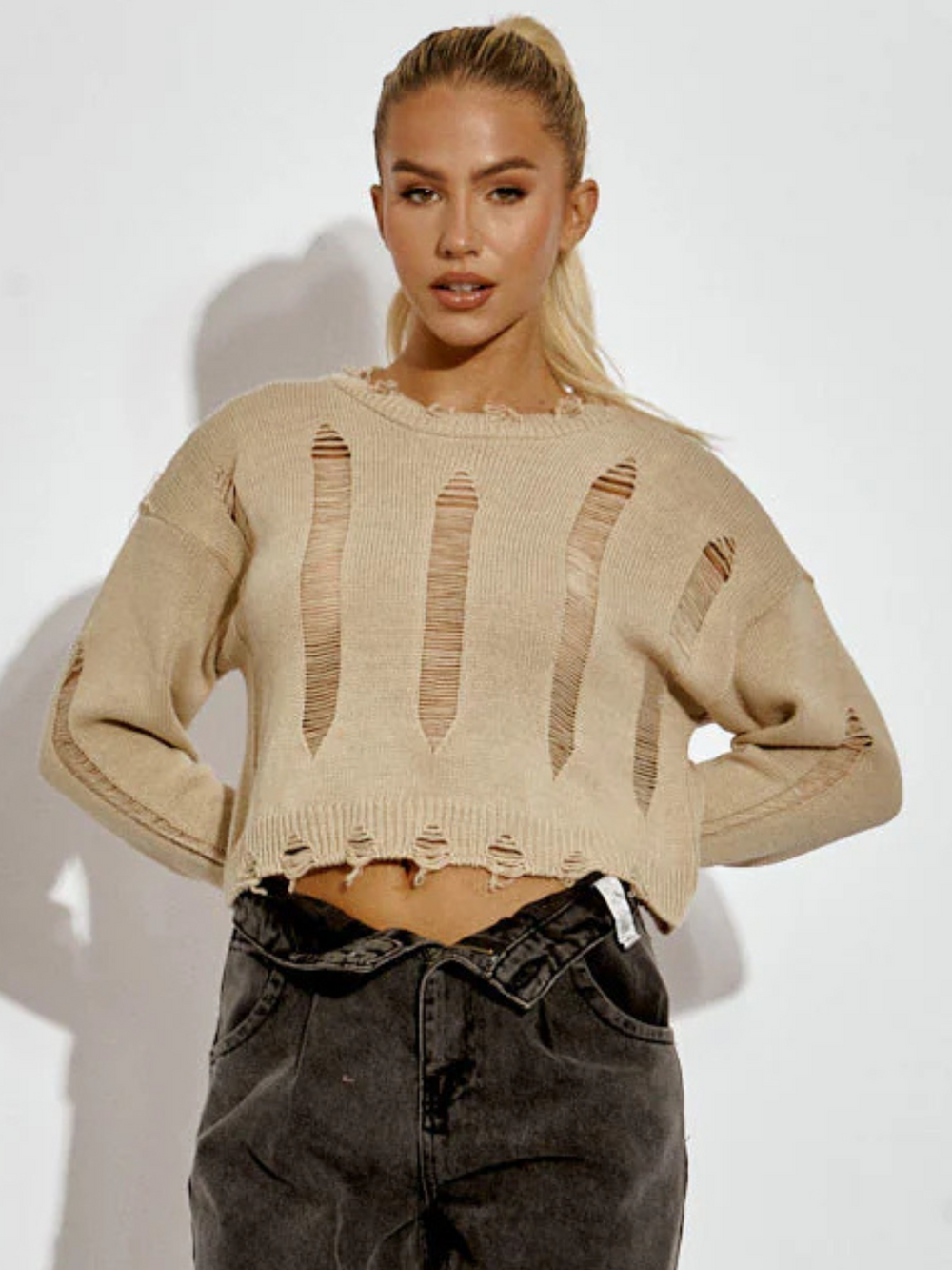 A female model wearing a stone long sleeve distressed style knitted cropped jumper in stone color paired with black jeans. The stone distressed knitted jumper has distressed panelling  and distressed hems. The model has tanned skin and long blonde hair that is in a ponytail.  The model faces the camera and places her hands behind he back.