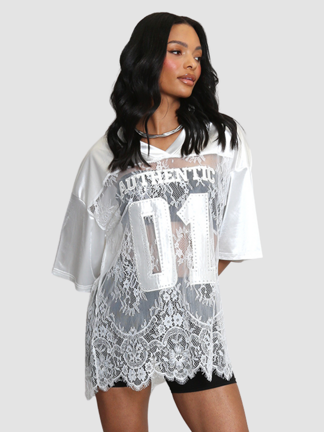 Female model wears a white lace varsity top with  with 3/4 satin sleeves. The varsity top has a sheer top with kace panel inserts. The model has light brown skin and long black hair, she looks off to the side. 