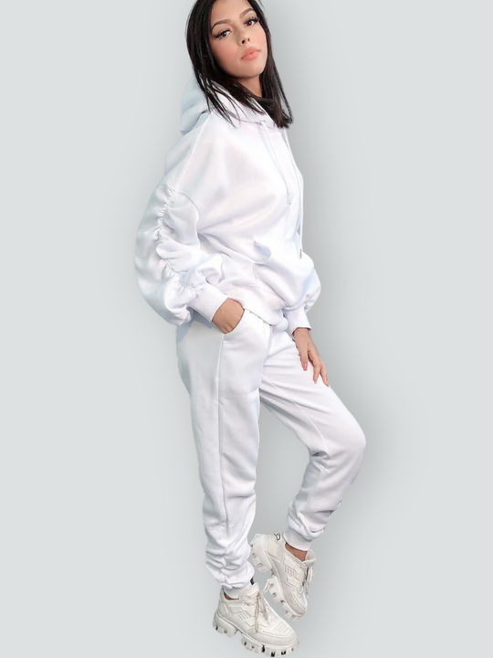 Full length shot. Model wears a ruched sleeve hooded jumper which has an adjustable hooded draw string  tightening if required. The hood comes with a matching white joggers that has a ruched design at the ankles. The model stands to the side, she turns her head slightly to face the camera and slightly tilts her head. Model stands to the side and tilts her head to the camera, she places one hand in her pocket and the other on her thigh. 