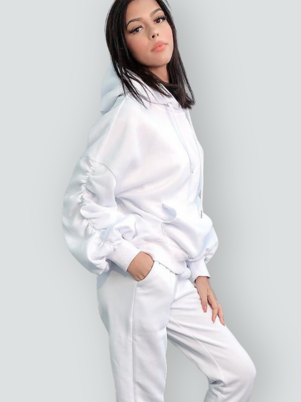 Model wears a ruched sleeve hooded jumper which has an adjustable hooded draw string  tightening if required. The hood comes with a matching white joggers that has a ruched design at the ankles. The model stands to the side, she turns her head slightly to face the camera and slightly tilts her head. Model places one hand in her pockets and the other on her thigh. 