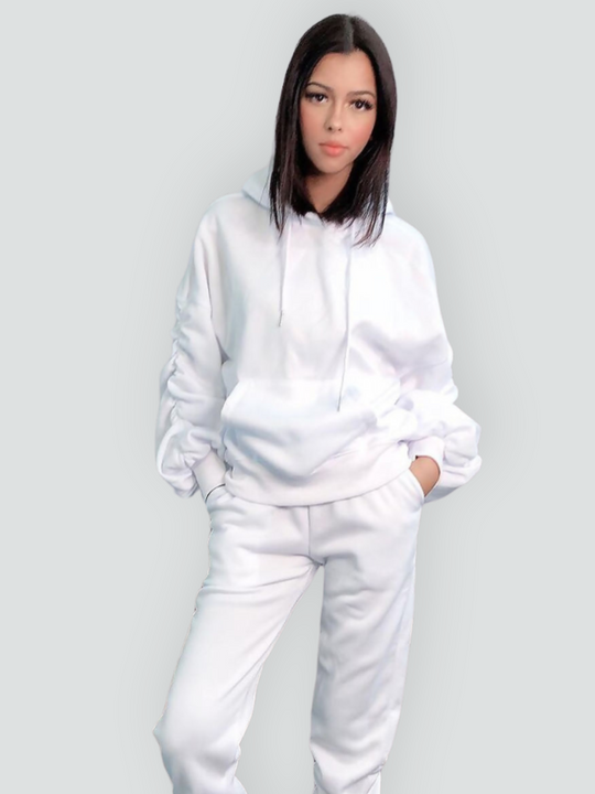 Mid length shot. Model wears a ruched sleeve hooded jumper which has an adjustable hooded draw string  tightening if required. The hood comes with a matching white joggers that has a ruched design at the ankles. The model stands to the side, she turns her head slightly to face the camera and slightly tilts her head. Model faces the camera and places both hands in her jogger pockets. 