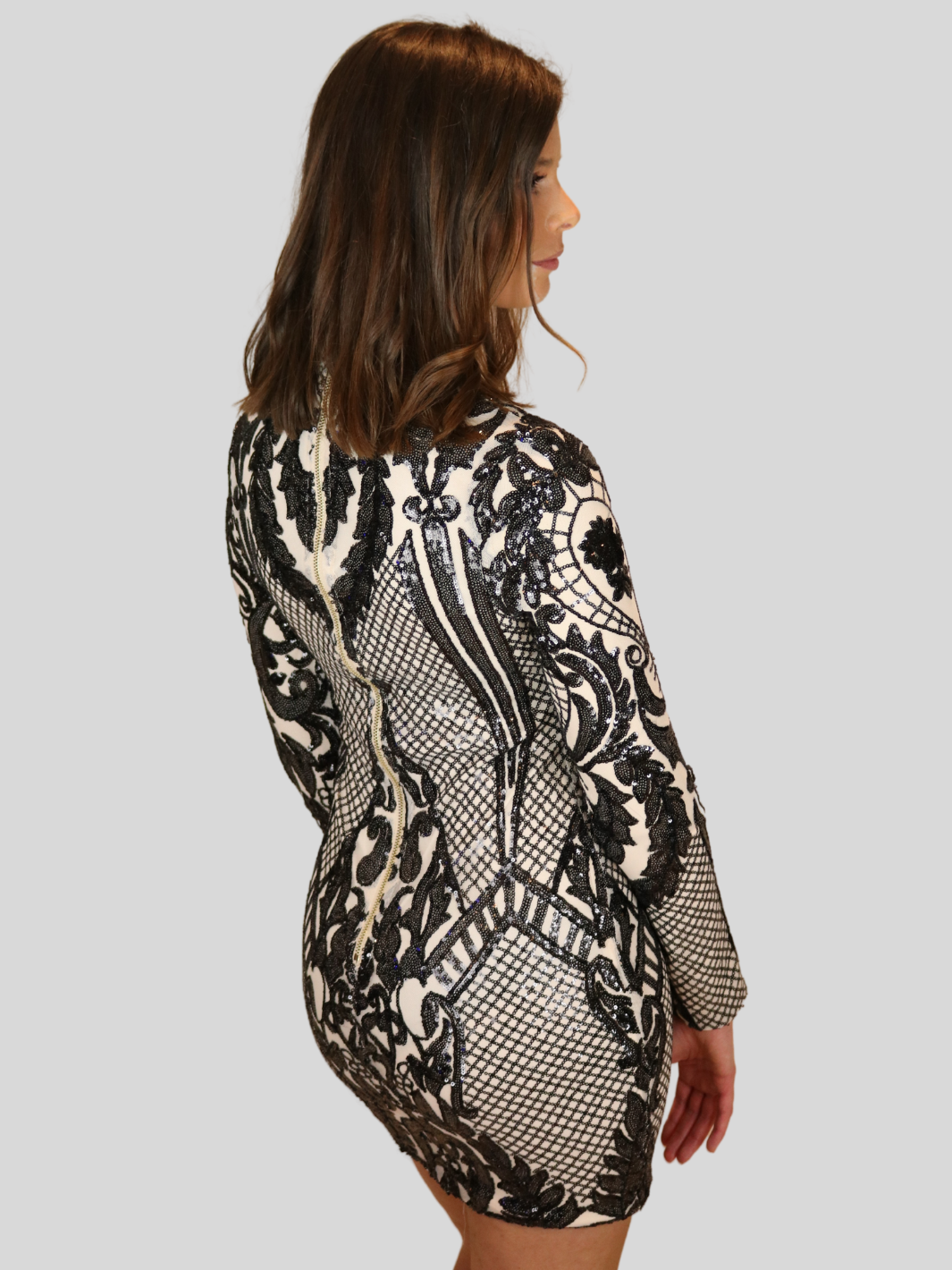 Model wears 4ever stunning premium deluxe a sequined  cream and black long sleeved dress, with a high neck and chunky gold back zip fastening. mModel has her back to the camera, the back of the dress is visible, showing the chunky zip fastening and gols and cream sequin design. 