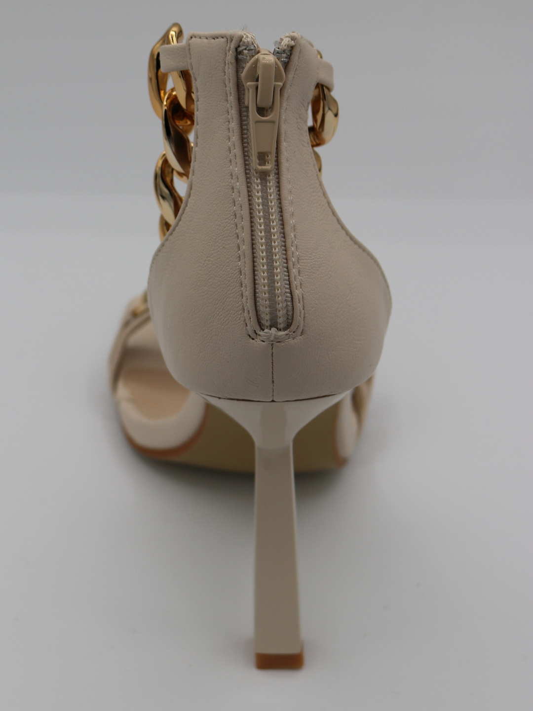 A cream shoe with gold detail at the base strap as well as gold chain detailing at the ankle. The photograph shows a close up of the back of the shoe, showing the cream heel and zip to the back of the shoe, that closes and secures the shoe. 