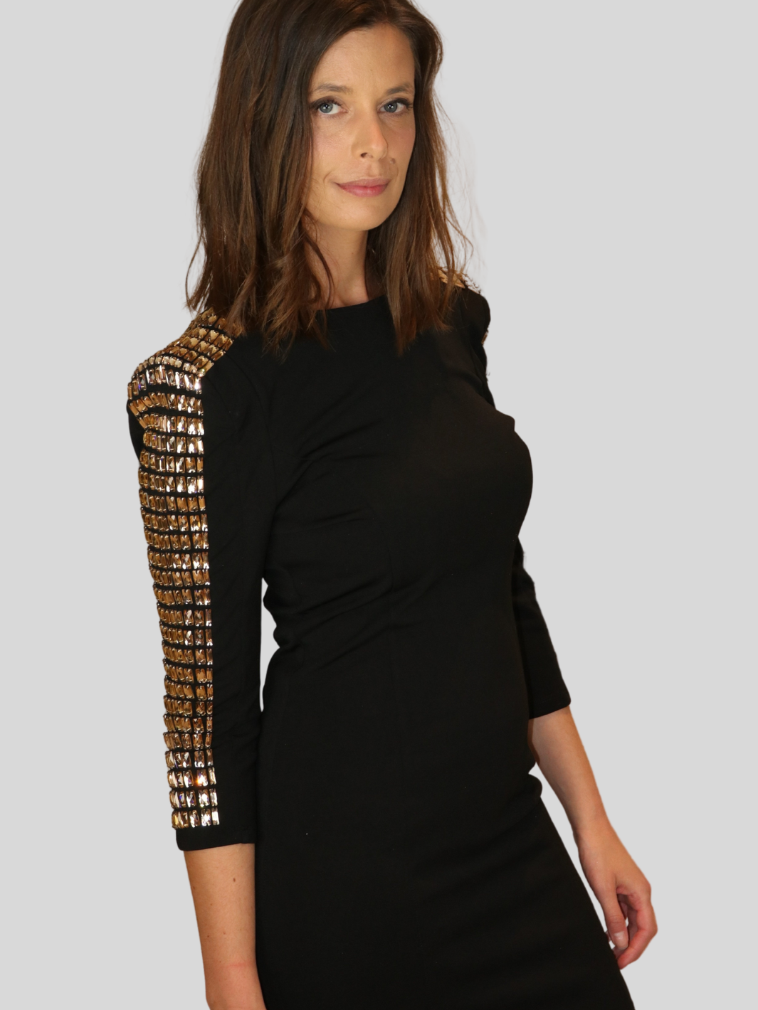 4ever Stunning Deluxe Premium black midi dress with gold rhinestone jeweled arms and chunky gold back zip fastening. Model stands to the side with her rihnestone jeweled sleeve visible. Model turns her head so that she is looking directly into the camera.