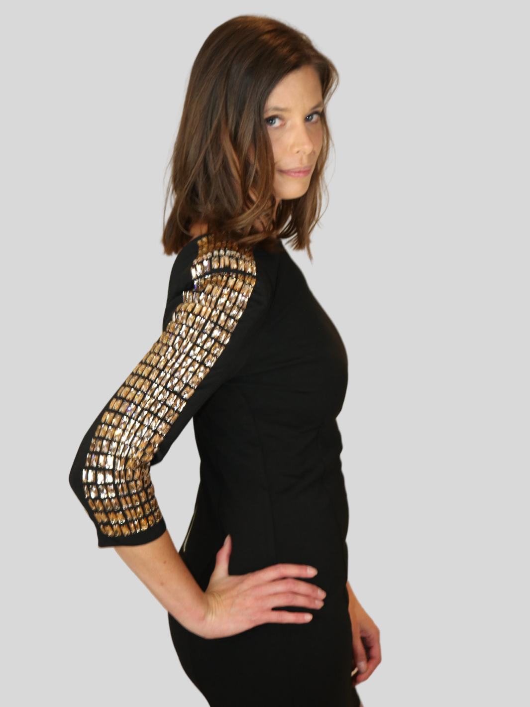 4ever Stunning Deluxe Premium black midi dress with gold rhinestone jeweled arms and chunky gold back zip fastening. Model stands to the side with her rihnestone jeweled sleeve visible. Model turns her head so that she is looking into the camera.