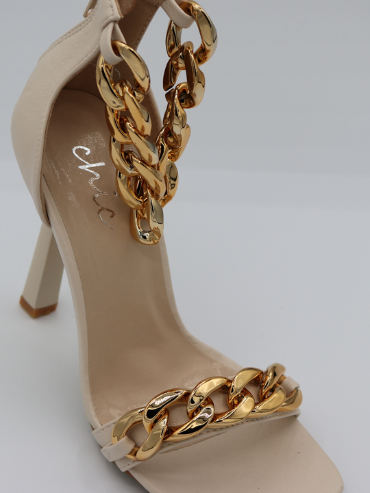 A cream shoe with gold detail at the base strap as well as gold chain detailing at the ankle. Shoe placed slightly to the side so that the front and side of the shoe can be seen.