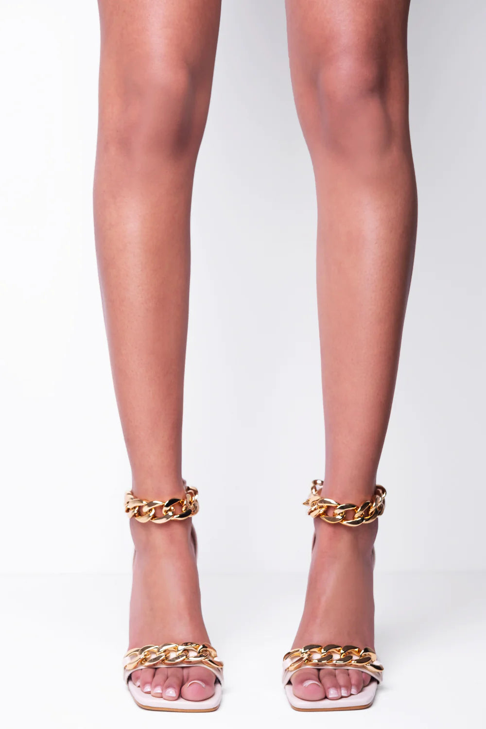 Model wears a cream shoe with gold detail at the base strap as well as gold chain detailing at the ankle. Model faces forward. The chunky gold detailing can be seen around the ankle and  as a strap that goes across the mid point of the shoe. 