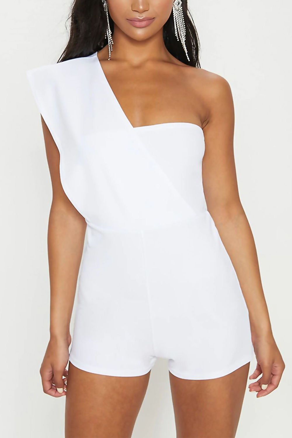 Model wears a white drape one shoulder playsuit. Model faces the camera and has both hands by her side. Model wears silver drape earrings. 