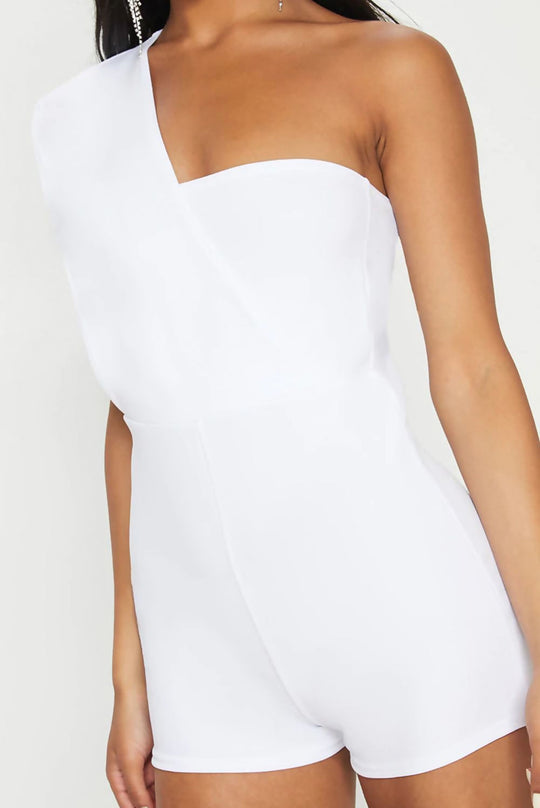 Model wears a white drape one shoulder playsuit. Model stands slightly to the side, where you can see one exposed shoulder and the other shoulder wearing a cross drapped sleeve. 