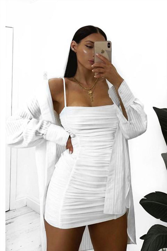 Model wears a white slinky ruched mini dress with spagehetti straps. Model wears and oversized shirt and holds her phone to take a selfie. 