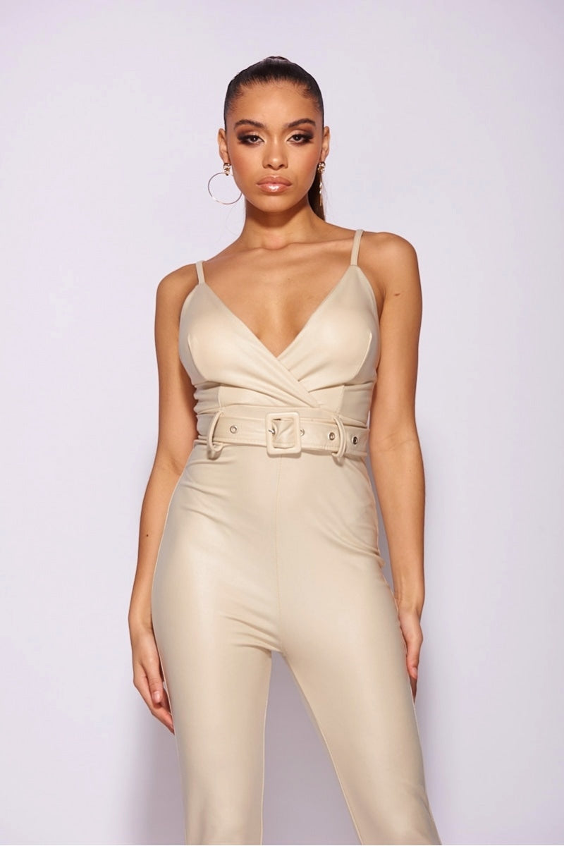 Model wears faux leather beige jumpsuit with high waisted belt . The jumpsuit has a skinny fit  style and a plunging neck line.  Model stands with her hands to the side and steers straight into the camera. 
