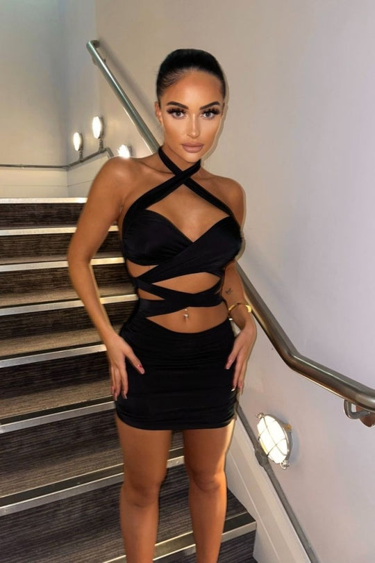 Black slinky bodycon dress with cut out mid section with cross over straps. The dress has a multiway function, you can wear it as a halter neck cross strap or as a standard halterneck. Model wears the halterneck as a cross neck halter. Model stand on the stairs and has both hands on her upper thighs. Model stares directly into the camera. 