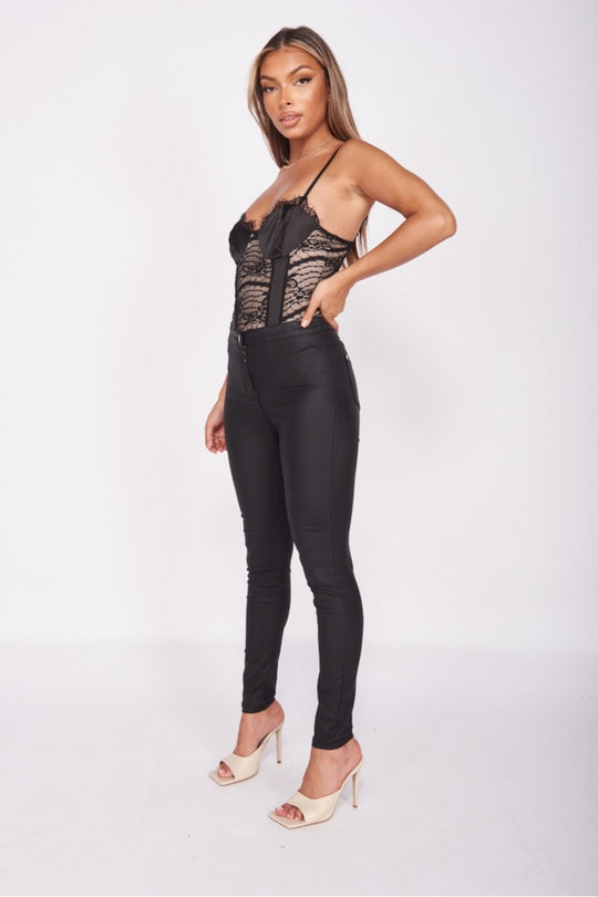 Black and tanned Body suit with eye lash lace trimmed detail and satin panneling. Model facing facing to the side with her hands on her hips.