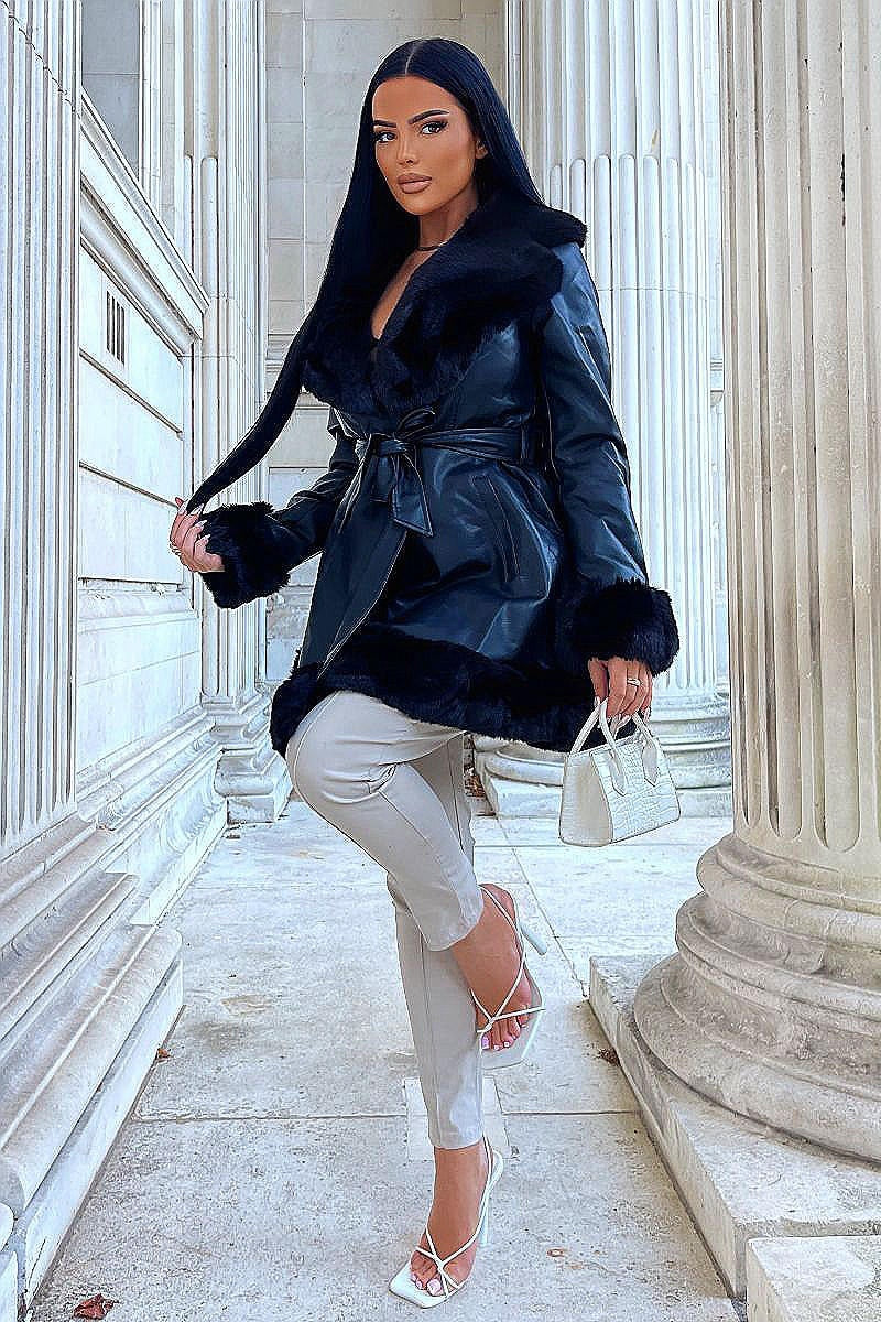 Model wears faux leather swing coat with faux fur tirm at the color, sleeve cuff and the bottom of the coat. The coat has a central button fastening and a faux leather belt. Model  is outside. Model raises one her leg, and holds a small bag in one hand and holds  her hair in the other.