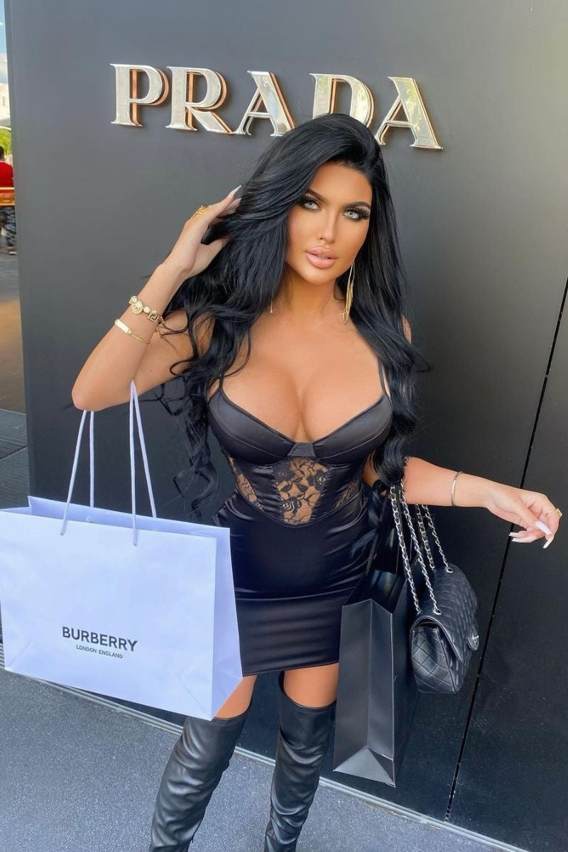 Model wears a black satin mini dress with cup detailing and front lace panelling. The dress has spaghetti straps and a concealed side closure.  Model stands outside holding shopping bags. Model places one hand in her hair. 