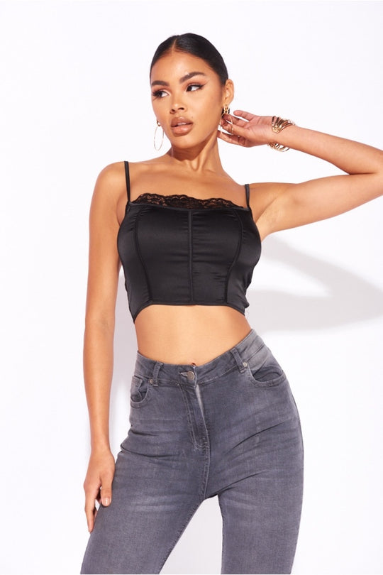 Model wears a black satin crop top with top lace detailing. Model wears grey skinny fit jeans.  Model has one hand behind her head and one at her side. Model looks to the side. 