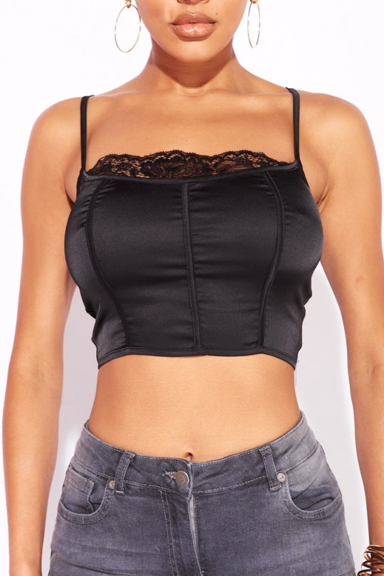 Model wears a black satin crop top with top lace detailing. Model wears grey skinny fit jeans. Model stands with both hands at her sides. 