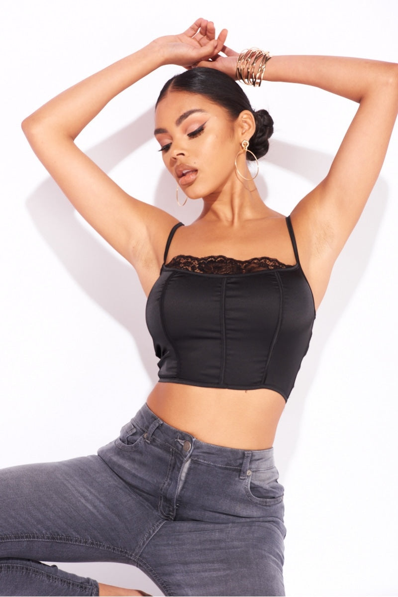 Model wears a black satin crop top with top lace detailing. Model raises her hands above her head, and looks down. Model wears grey skinny fit jeans. 