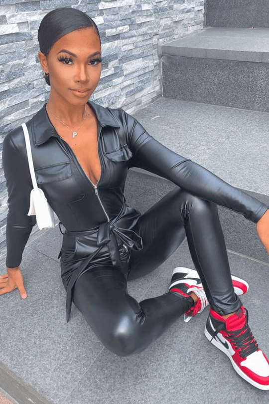 Model wears faux leather black jumpsuit that has long sleeves and front zip fastening. The jumpsuit has a faux leather removable belt at the waist. Model has the jumpsuit partially zipped so that some of her torso is visible. Model is outside. Model is sitting on a step and stares directly into the camera. Model wears red, white and black trainers. 