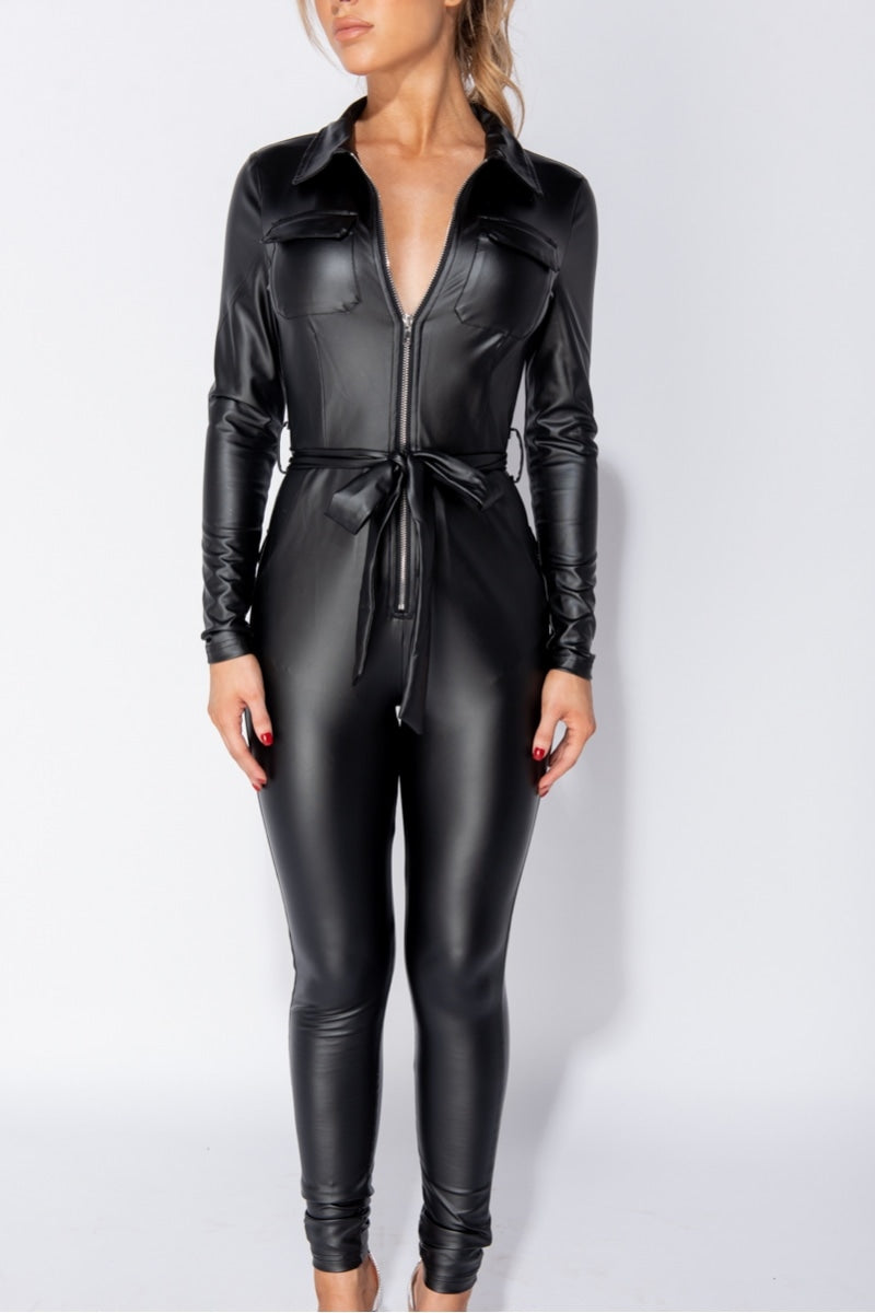 Model wears faux leather black jumpsuit that has long sleeves and front zip fastening. The jumpsuit has a faux leather removable belt at the waist. Model has the jumpsuit partially zipped so that some of her torso is visible. Model stands with her arms by her sides and looks to the side.