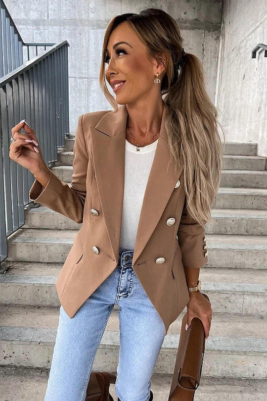 Model wears a camel coloured military button inspred blazer with padded shoulders and button fastening. Model stands on a step. She looks to the side whilst  smiling. Model holds a brown clutch bag and is wearing light blue denim jeans.
