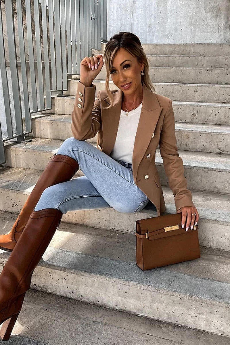Model wears a camel coloured military button inspred blazer with padded shoulders and button fastening. Model sits on a step and looks up. Model holds a brown clutch bag and matching shoes. Model  is wearing light blue denim jeans. 