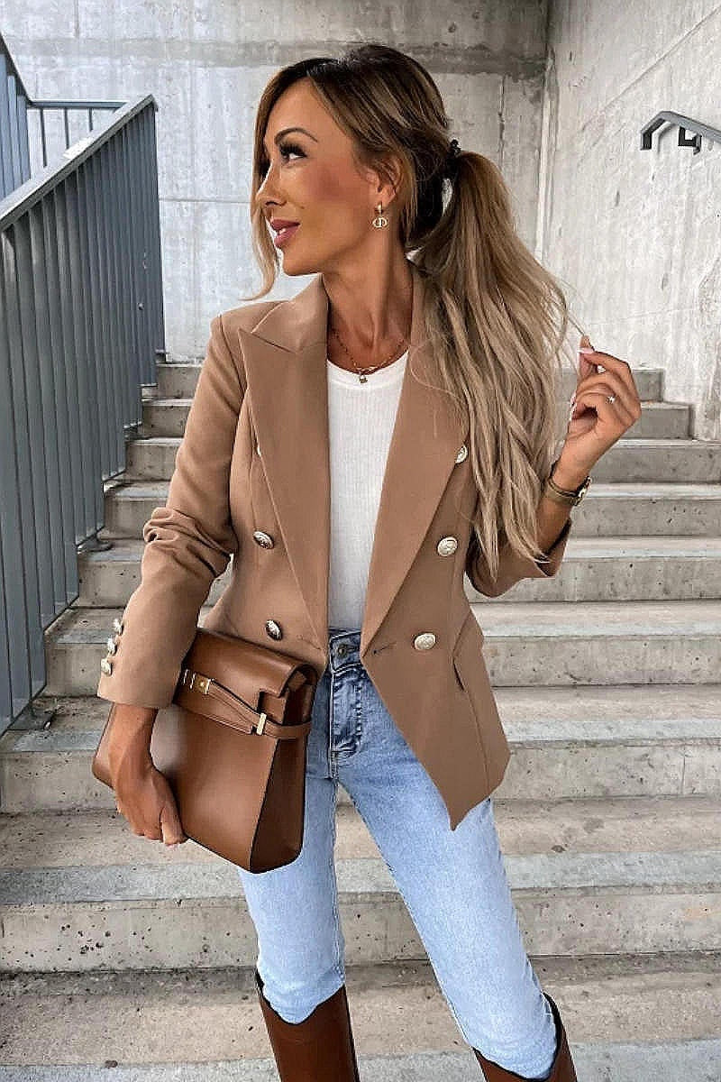 Model wears a camel coloured military button inspred blazer with padded shoulders and button fastening. Model's body  faces the camera wth one leg to te side and her head looking to the side. Model holds a brown bag and is wearingbrown boots. 