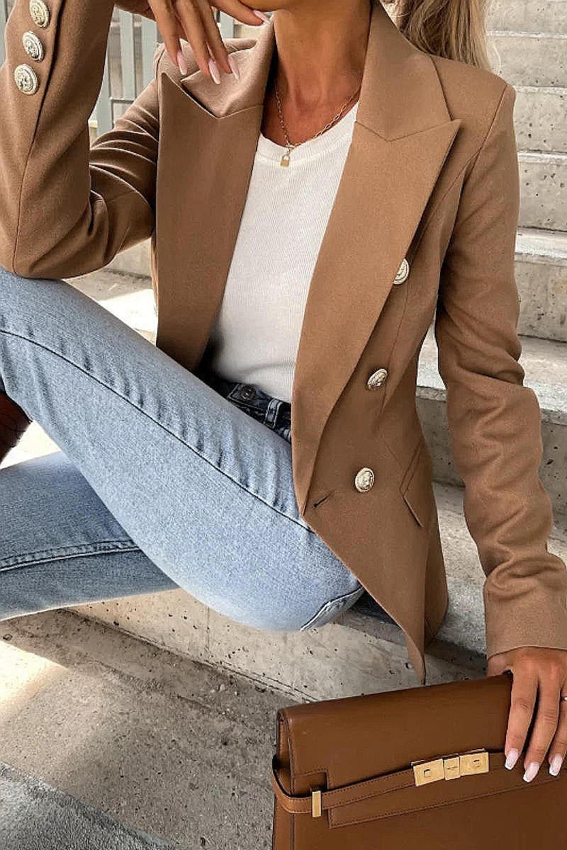 Model wears a camel coloured military button inspred blazer with padded shoulders and button fastening. Closeup of model sitting on a step with the blazer open and a brown clutch bag resting on the step. Model wears blue jeans.