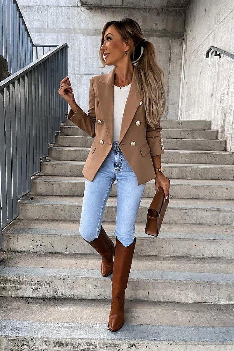Model wears a camel coloured military button inspred blazer with padded shoulders and button fastening. Model stands on a step. She looks to the side whilst smiling. Model holds a brown clutch bag and is wearing light blue denim jeans and brown matching boots.