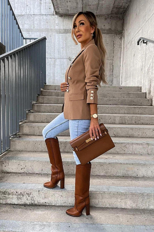 Model wears a camel coloured military button inspred blazer with padded shoulders and button fastening. Model stands on the step and looks off into the distance. Model holds a brown clutch bag and wears matching brown boots.