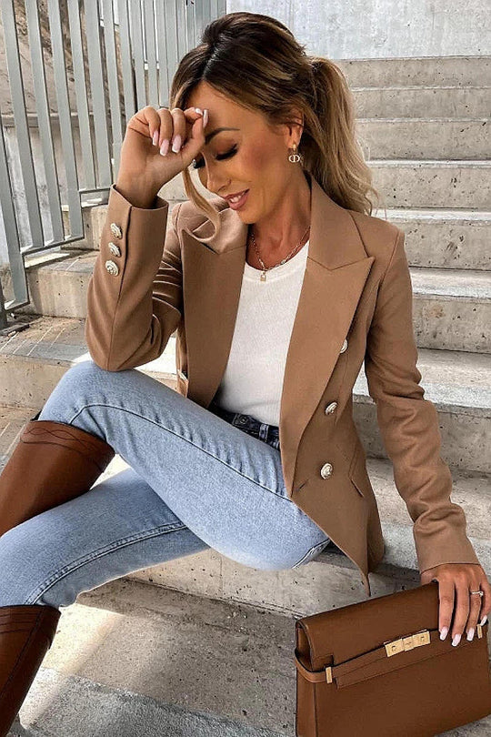 Model wears a camel coloured military button inspred blazer with padded shoulders and button fastening. Model sits on a step and looks down with one hand on her head.  Model holds a brown clutch bag and is wearing light blue denim jeans. Model