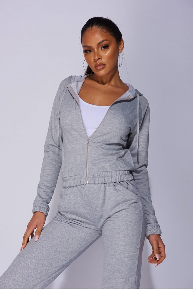 Model wears a light weight tracksuit with front zip fastening and draws string hood.  Model faces the camera with one leg to the side. 