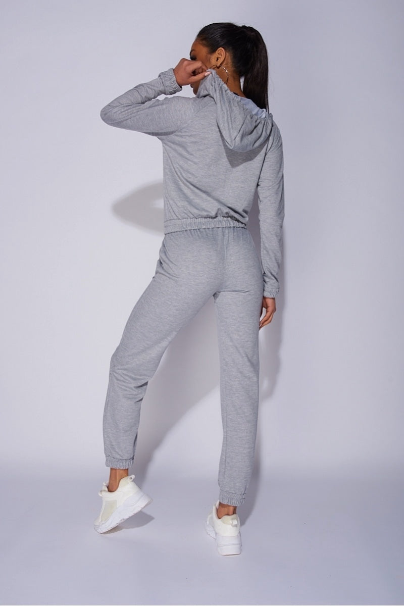 Model wears a light weight tracksuit with front zip fastening and draws string hood. Model places her back too the camera. The back of the tracksuit is visible, including the hood which the model holds with one hand. 