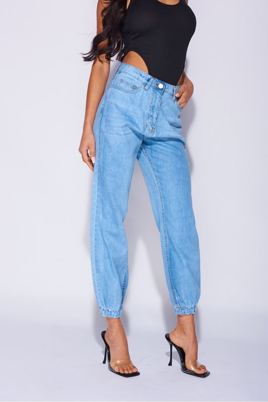 Model wears blue ciffed denim joggers with front zip fastening. Model wears a black high rise body suit. Model stands to the side. the front of the jeans is visible. 