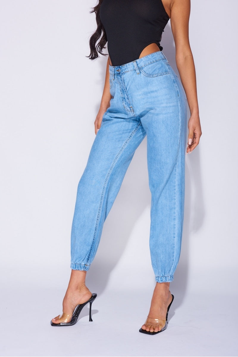 Model wears blue ciffed denim joggers with front zip fastening. Model wears a black high rise body suit.  Model stands to the side with one leg in front of the other and her hands by her sides. 