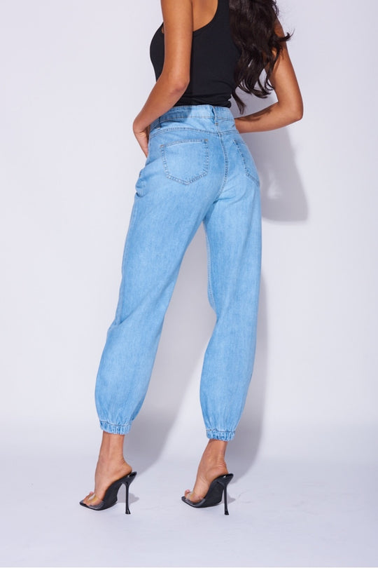 Model wears blue ciffed denim joggers with front zip fastening. Model wears a black high rise body suit. Model stands with her back to the camera, the back pocket detailing is visible. 