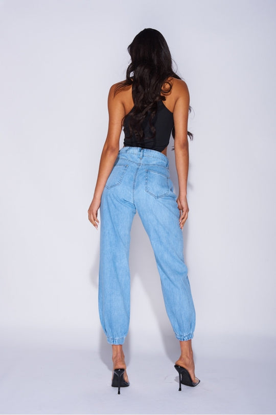 Model wears blue ciffed denim joggers with front zip fastening. Model wears a black high rise body suit. Model stands with her back to the camera. The back pocket detailing is visible. Model stands with both hands by her sides. 