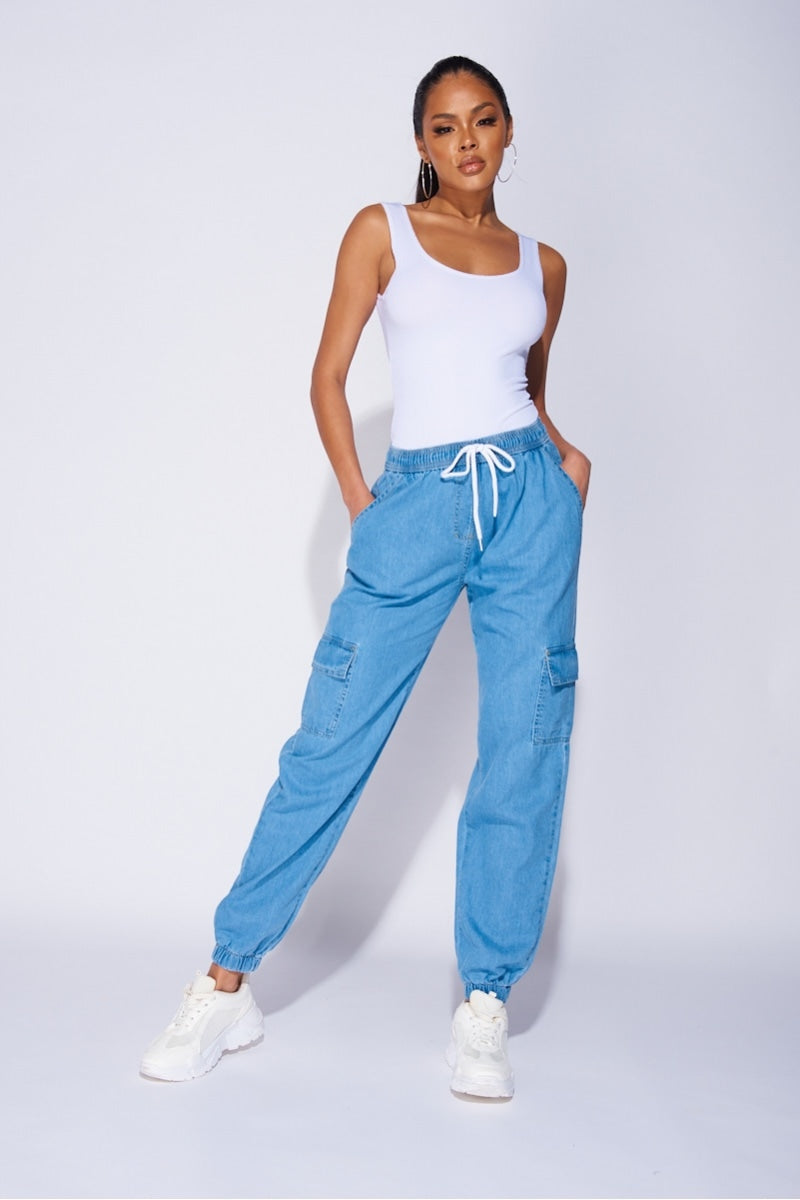 Model wears a loose fit light blue patch pocket cuffed denim joggers. Model wears a white bodysuit and white trainers.  Model stands facing forwars,slightly leaning to one side. 